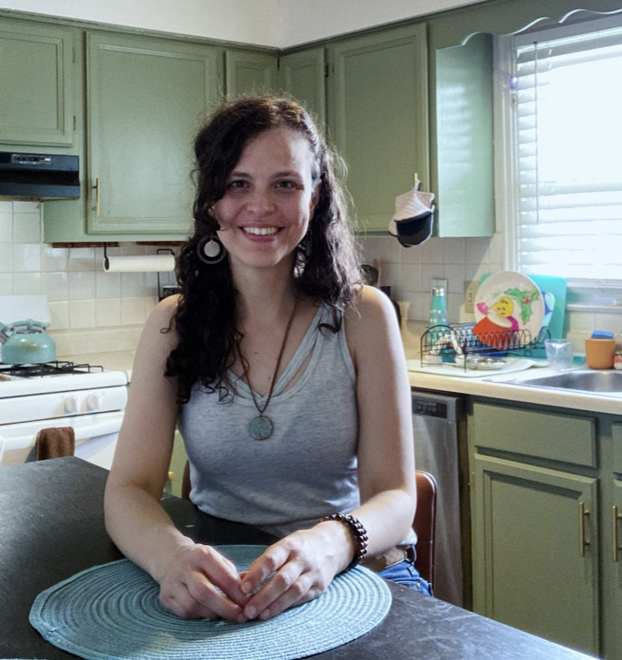 A woman with long, curly dark hair smiles while standing on the porch of a light-colored house
