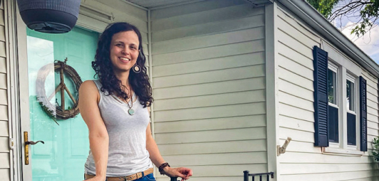 A woman with long, curly dark hair smiles while standing on the porch of a light-colored house