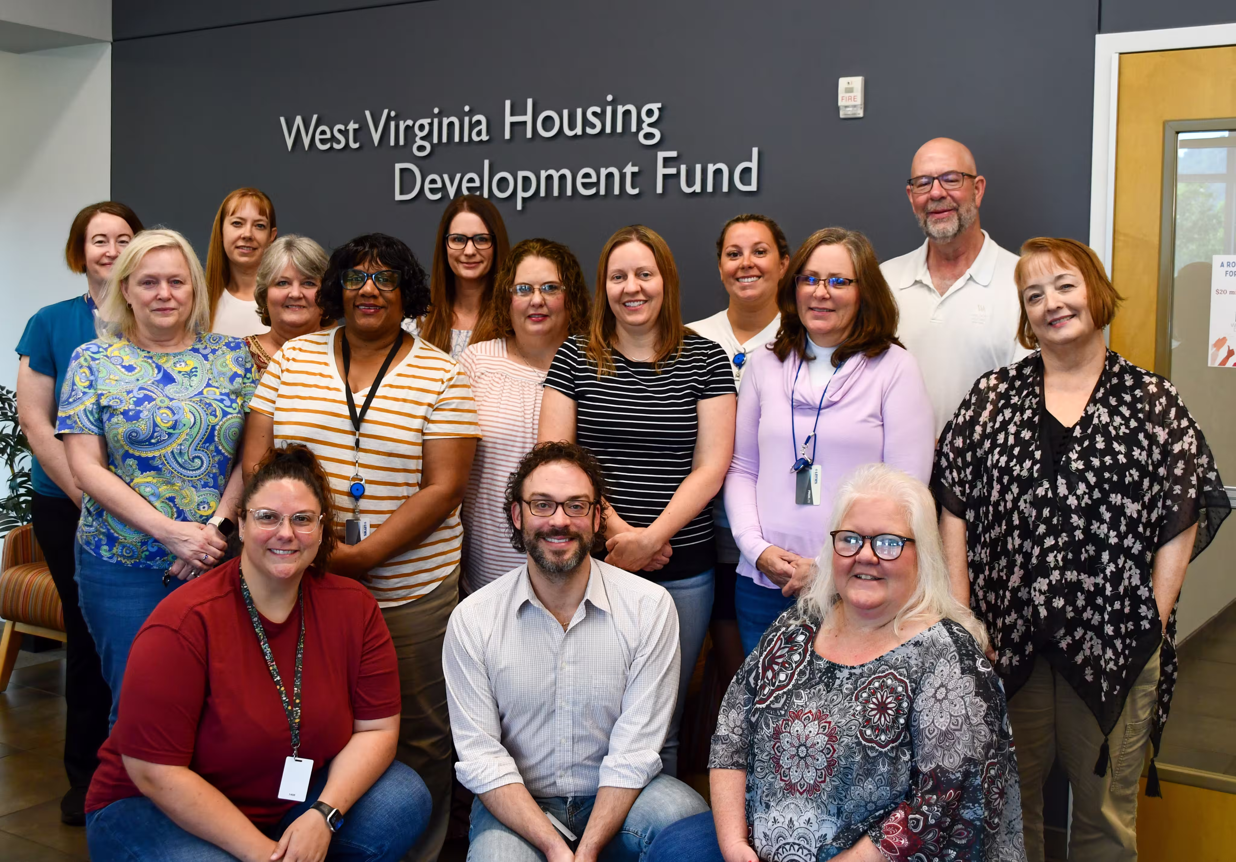 A group of fourteen people, diverse in age, gender, and ethnicity, pose for a team photo in front of a wall with the sign 'West Virginia Housing Development Fund.' They are dressed in a mix of casual and business casual attire, with some wearing ID badges. The group is smiling, standing and kneeling in two rows in an office-like setting with a door and a window in the background.