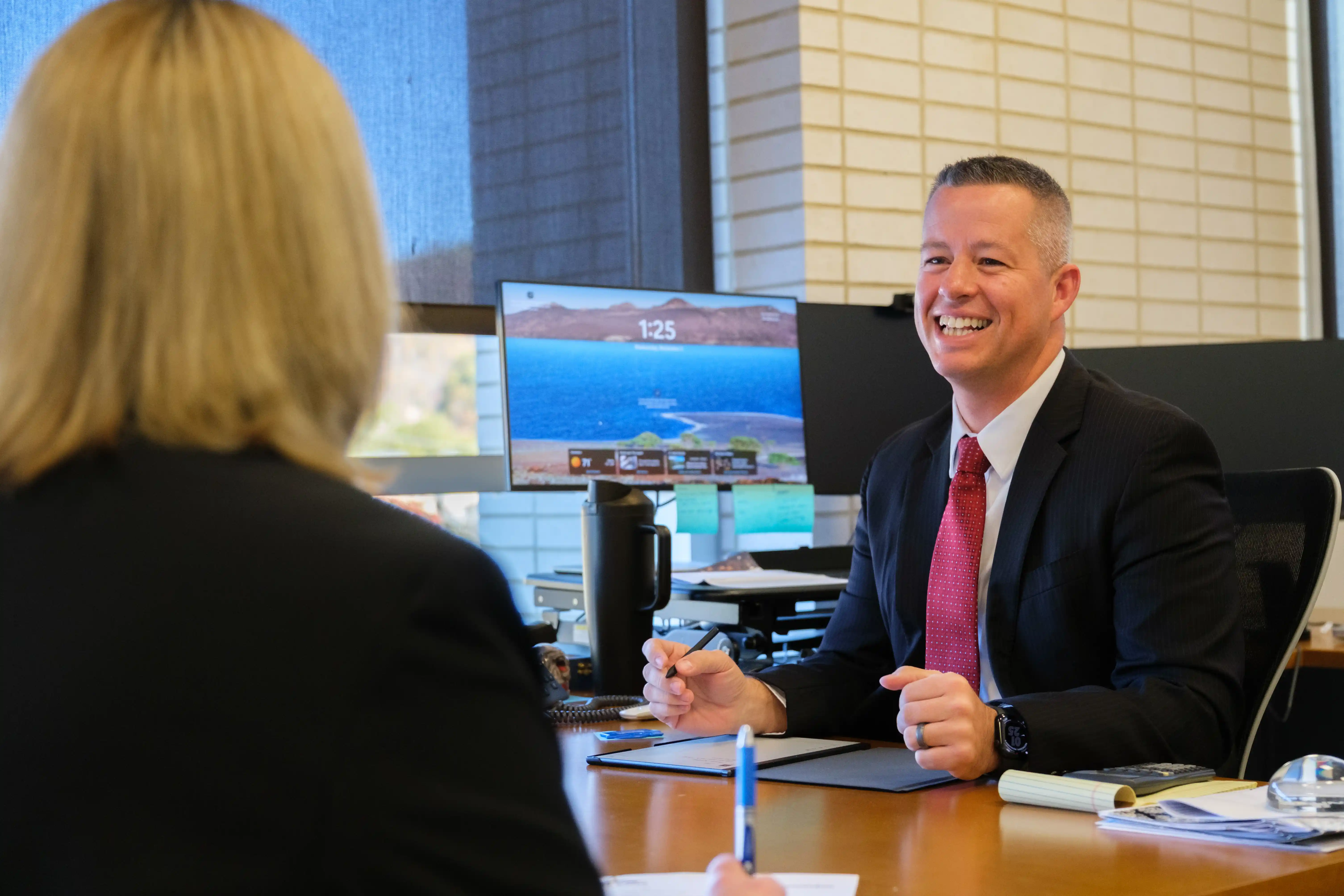 A smiling man in a suit sits at an office desk engaging in conversation with a colleague whose back is turned to the camera