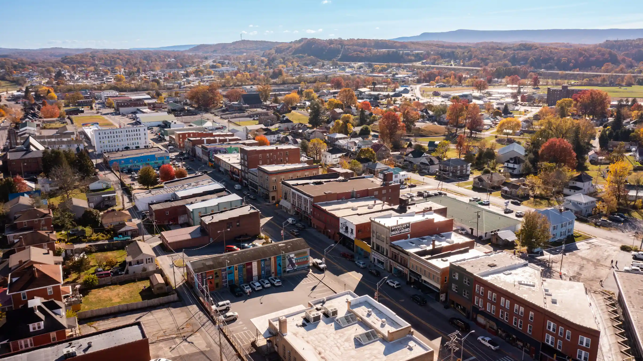 An aerial view of downtown Princeton, West Virginia, bathed in sunlight, featuring brick buildings, a colorful book-spine mural, and vibrant autumn foliage with rolling hills in the background.