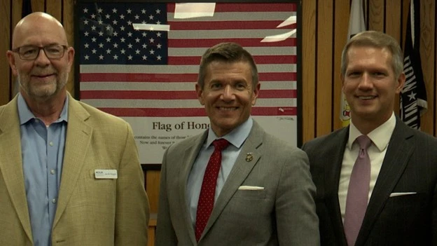 Three men in suits stand in front of an American flag.