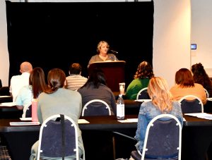 Kay Bowe, the Fund's Underwriting Manager, stands in front of a seated crowd at the Holiday Inn in South Charleston. She is standing in front of a black curtain.