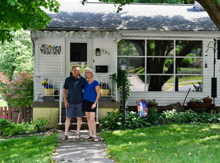 A man in a dark t-shirt and shorts stands next to a woman with a blue t-shirt and black shorts on a sidewalk in front of a small, white, single-story home.