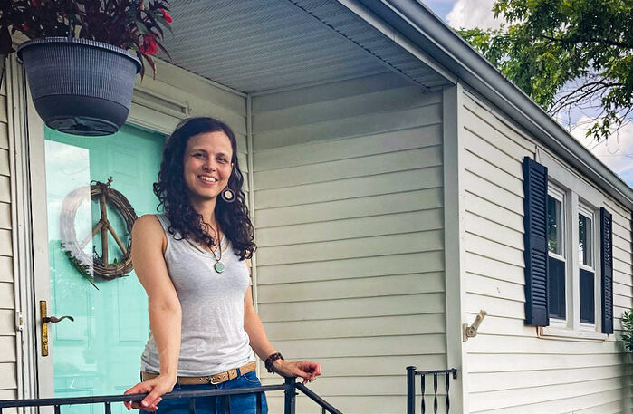 Young woman with brown hair wearing a white tank top and blue jeans stands on the front porch of her home with a blue front door in the background.