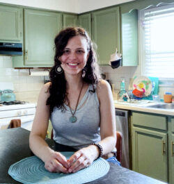 A young woman with long brown hair wearing a white tank top stands at her kitchen counter. Behind her are green cabinets.