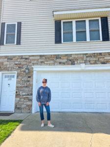 A woman in a gray sweatshirt and blue jeans stands in front of the white garage door to her townhome. The townhome is beige vinyl on top and various shades of beige and brown stone on bottom. 