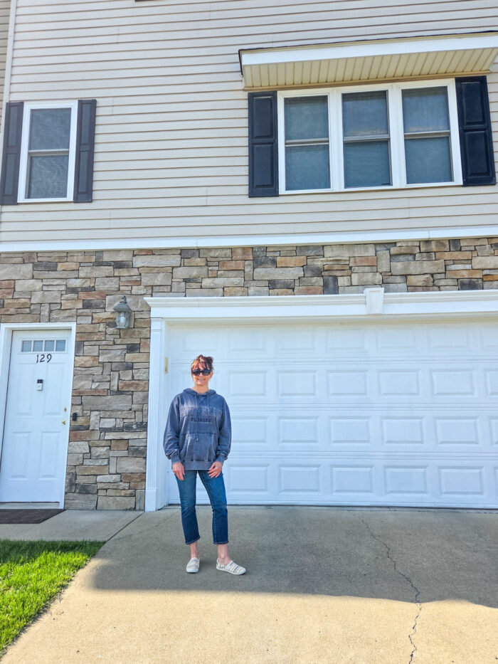 Person in casual clothes standing on driveway in front of a house with stone and beige siding and white garage door.