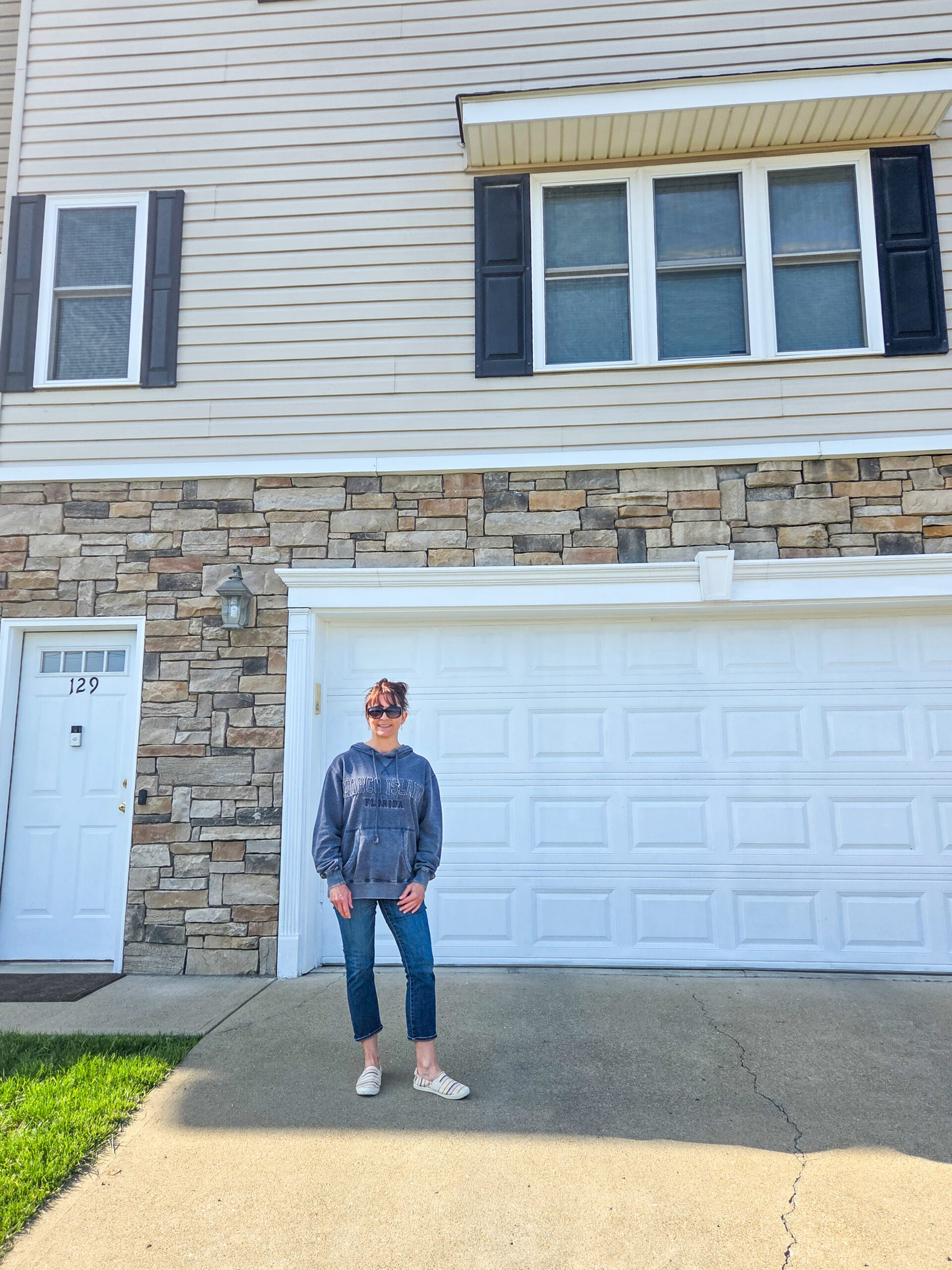 Person in casual clothes standing on driveway in front of a house with stone and beige siding and white garage door.