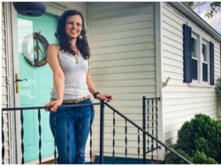 A young woman with long brown hair wearing a white tank top and dark denim jeans stands on her front porch. Her home is white vinyl siding with a light blue front door and black iron railings around the porch.