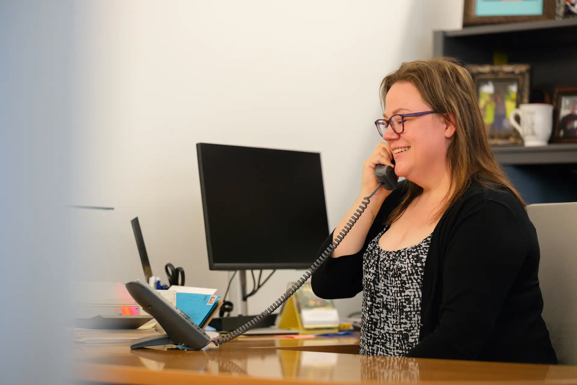 Person smiles while speaking on the phone at a desk, reflecting a helpful and approachable office environment.