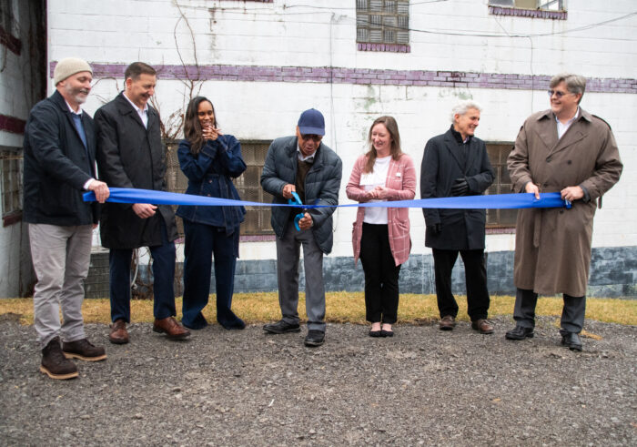 A group of people representing the City of Bluefield, the Bluefield Arts and Revitalization Coalition, and funding partners hold a blue ribbon as the man in the center cuts it with scissors. They are standing in front of the Hotel Thelma, an aging structure built of white cinder block. 