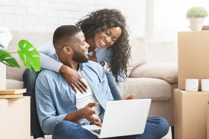 A smiling couple sitting on the floor using a laptop together surrounded by moving boxes.