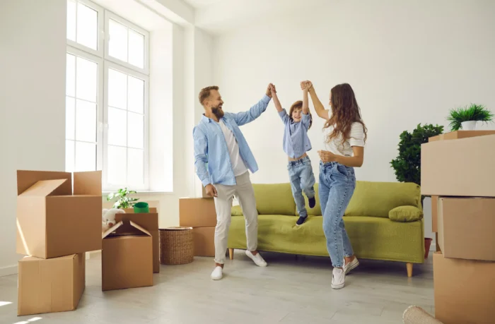 Parents holding hands with their young son as he jumps for joy in a living room filled with moving boxes.