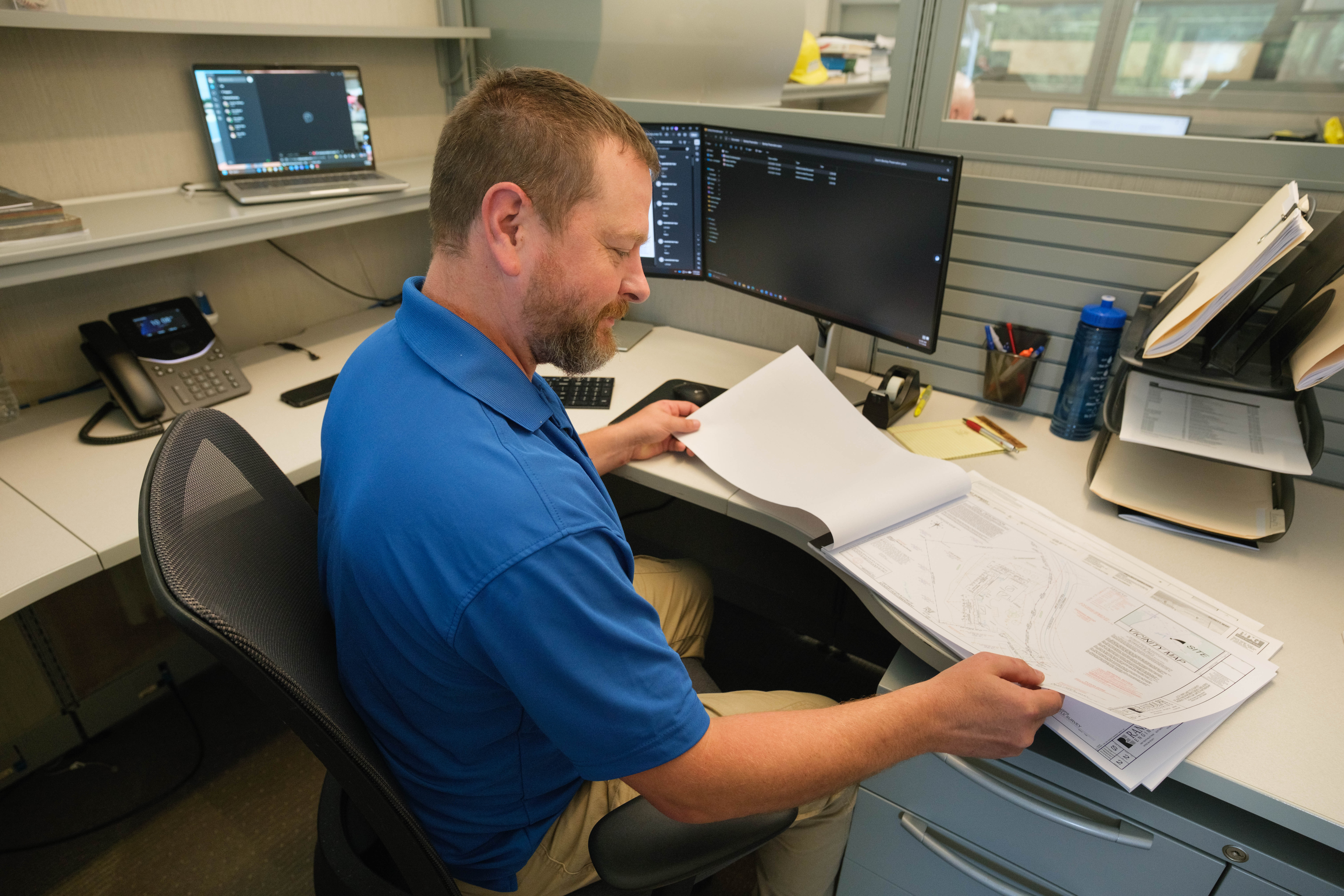 A man in a blue shirt sits at a work station and looks over building plan.